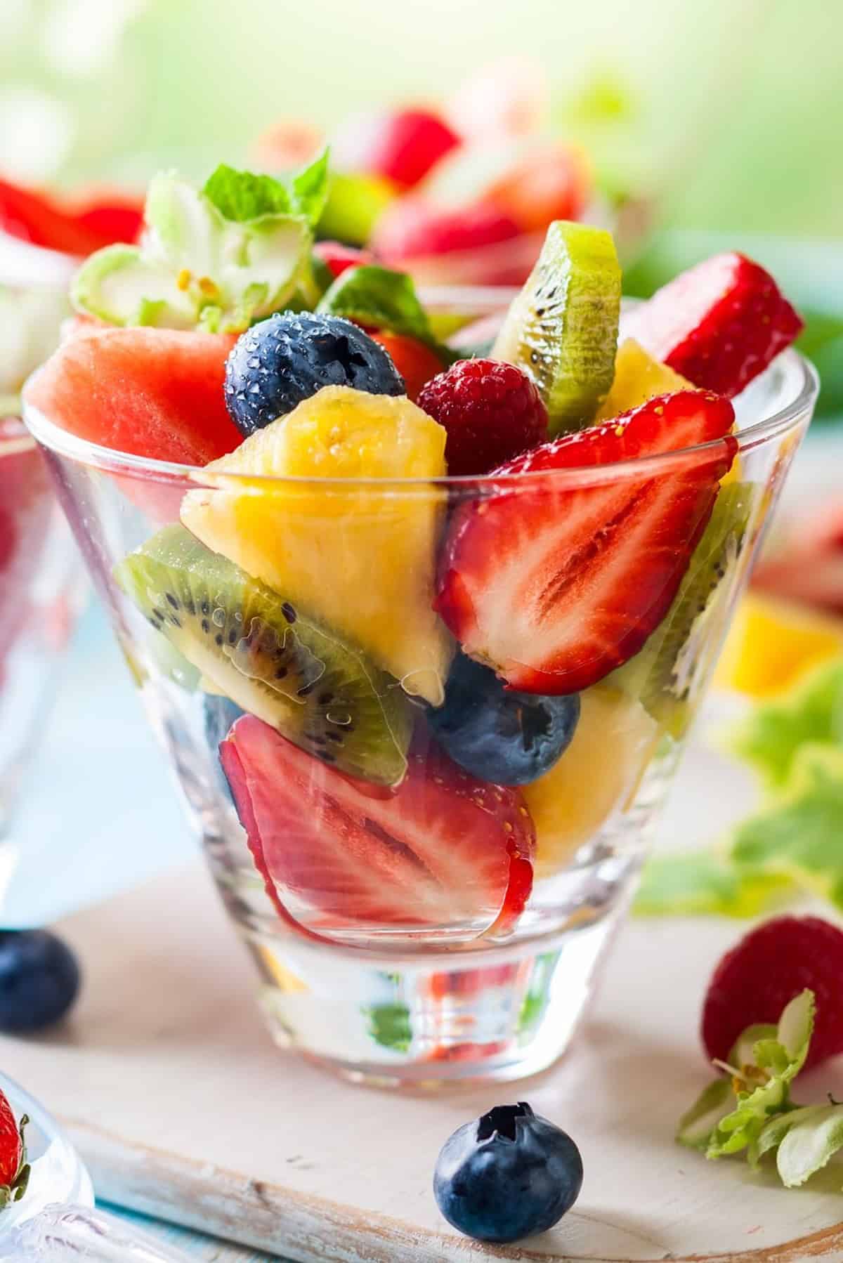 Tropical fruit salad in a glass cup with fresh fruits on a cutting board.