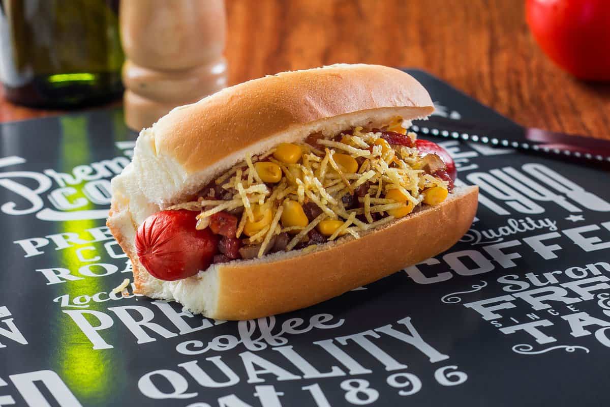 Brazilian hot dog sitting on a table with ingredients around it. 