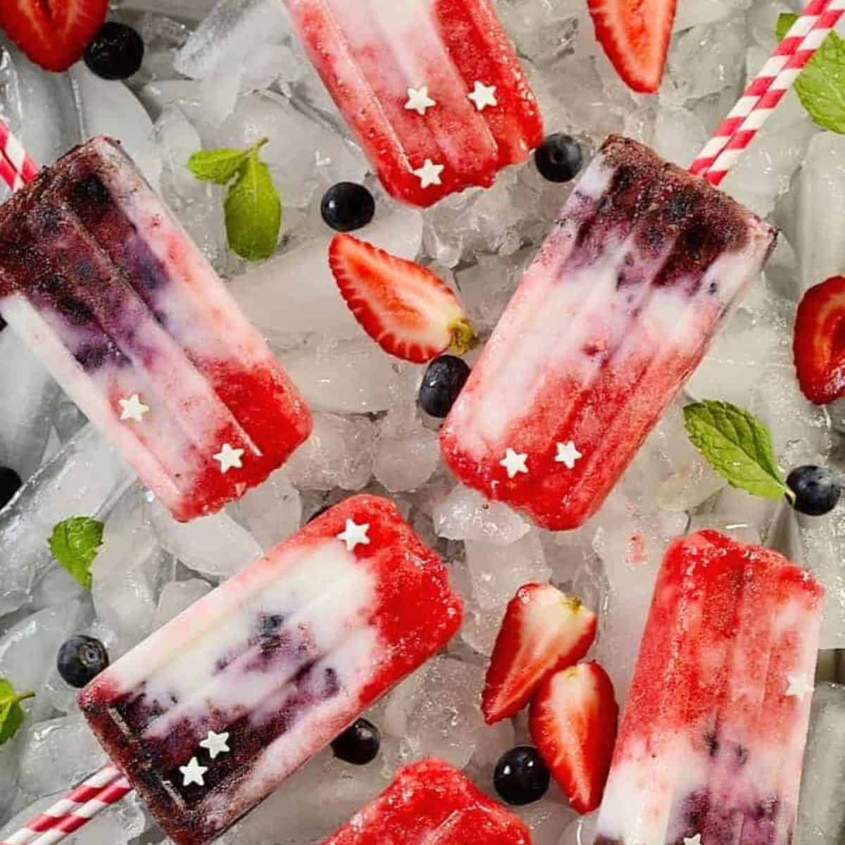 Several red, white, and blue berry yogurt ice pops on ice cubes with some scattered ingredients around them.
